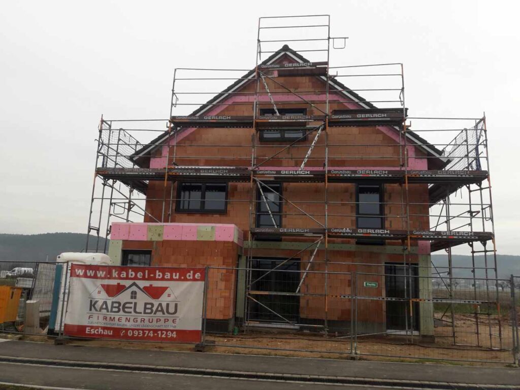 Frontansicht mit Gerüst und Banner; Dachdeckung nahezu fertig.