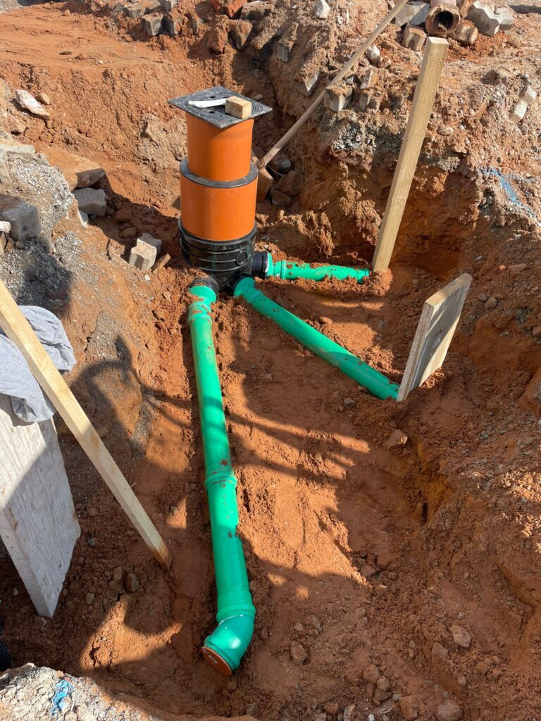 Grüne Abwasser-/Versorgungsrohre an orangefarbenem Kontrollschacht im Erdreich.