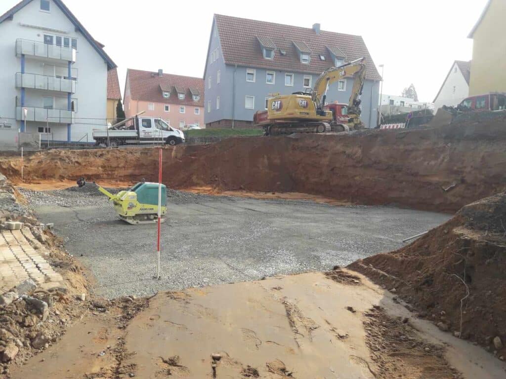Tiefe Baugrube mit Schotterplanum; Minibagger und Messlatte im Aushub.