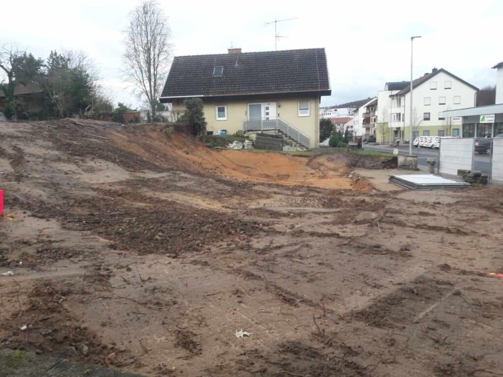 Geräumtes Baufeld mit Erdarbeiten; Boden abgetragen, Nachbarhäuser im Hintergrund.